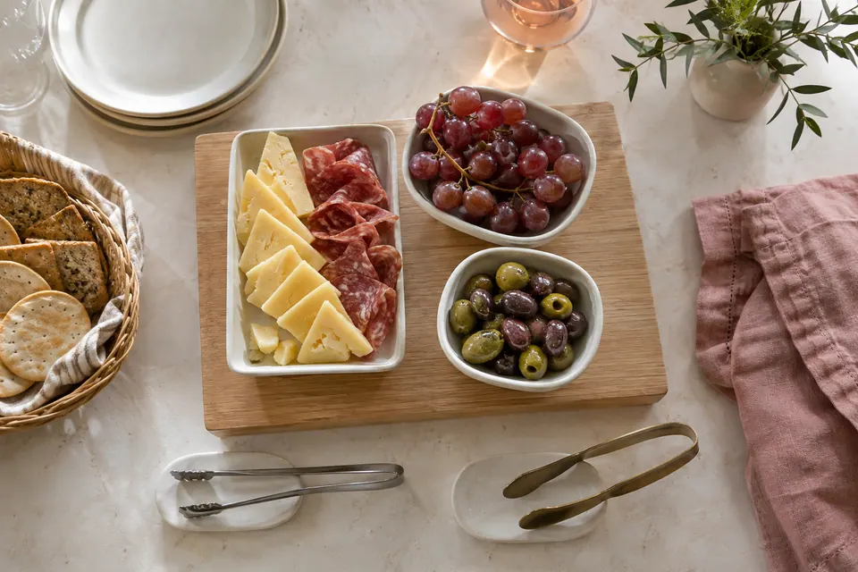 Cheese and salami share an ivory platter on an oak board, with separate grape and olive bowls and a cracker basket beside it.